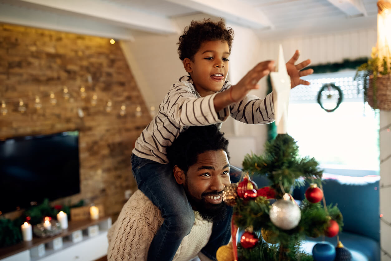 A black boy sitting on his fathers shoulders to put a star on top of a Christmas tree.