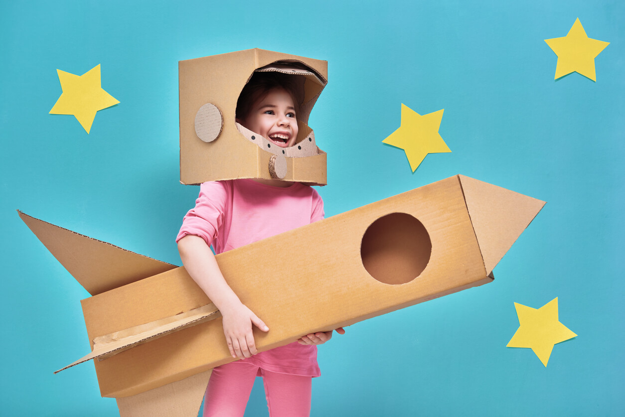A young girl wearing a carboard helmet and carrying a rocket made with cardboard boxes.