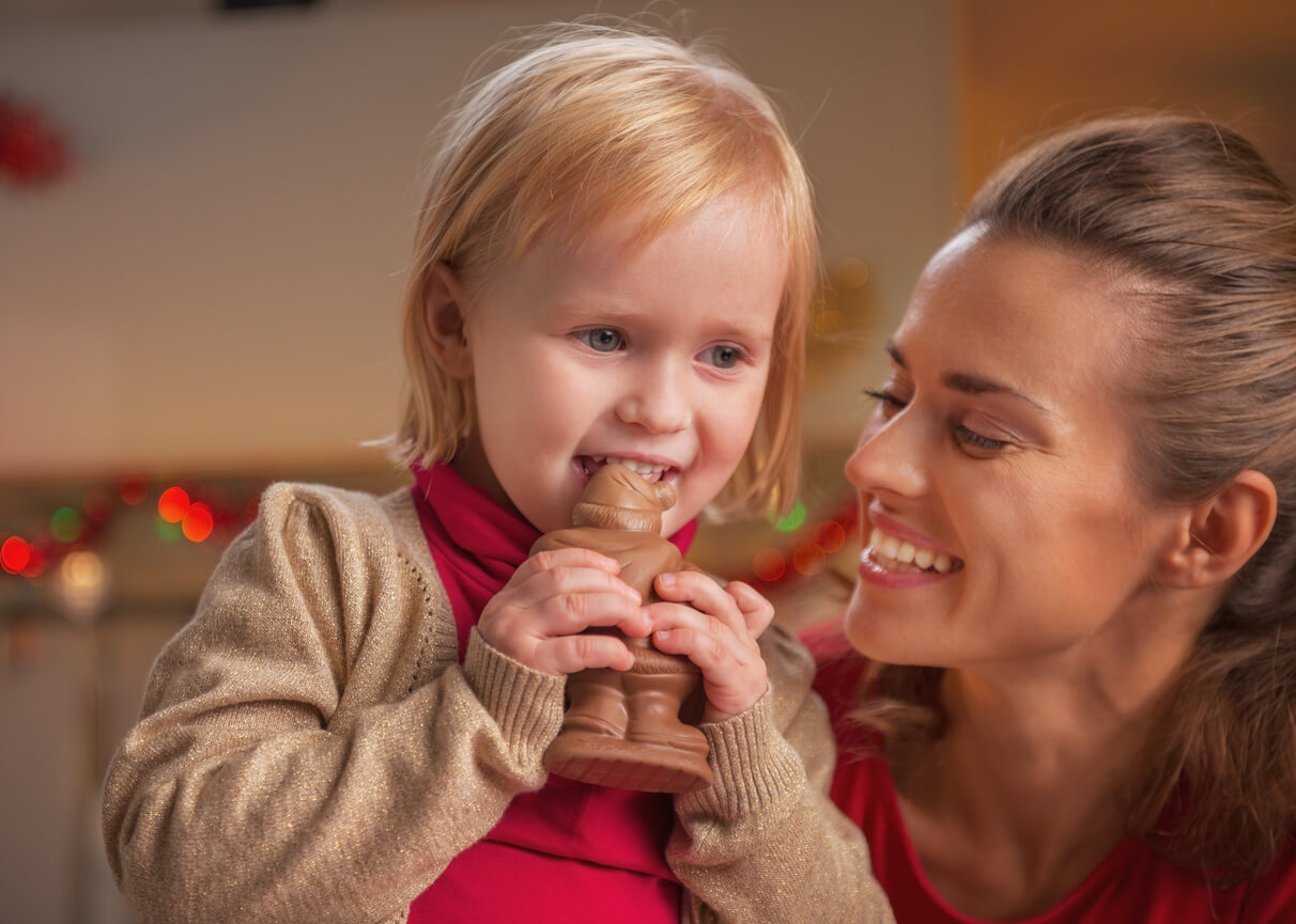A mother kneeling down next to her toddler daughter, who's biting into a large Santa-shaped chocolate figure.