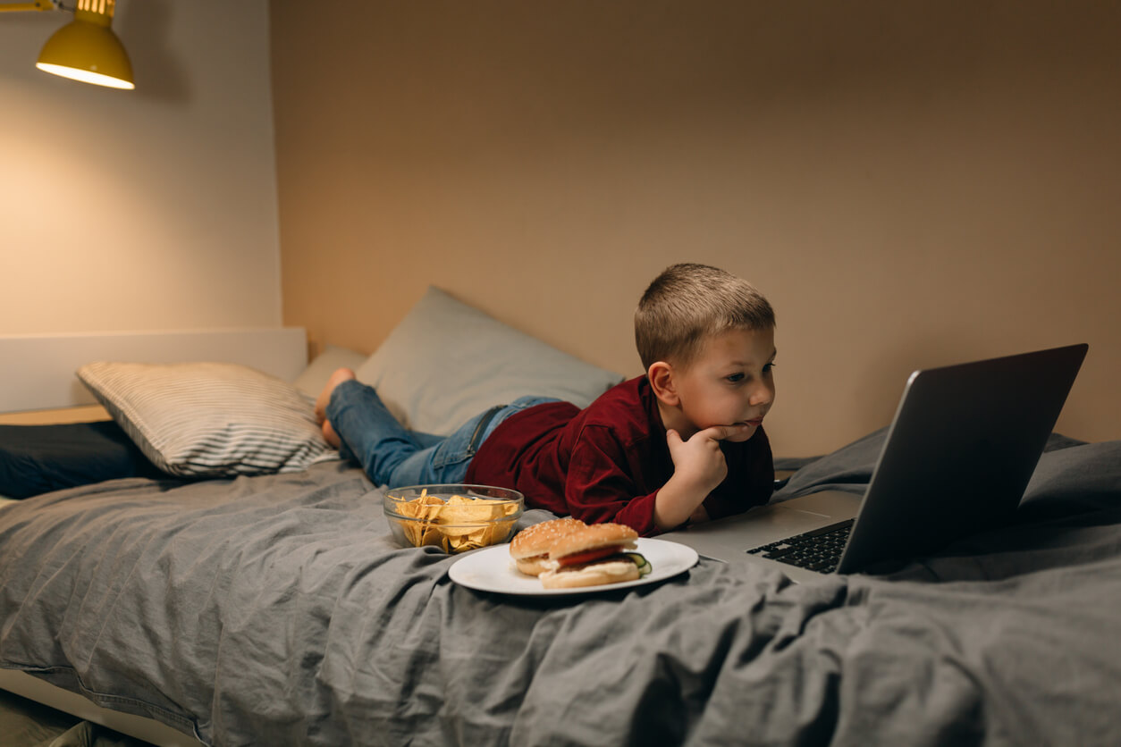 Ett barn som äter snabbmat medan han ligger i sängen och tittar på något på en bärbar dator.