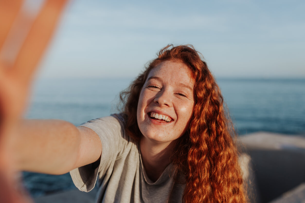 A red-headed teenage firl taking a natural looking selfie at the beach.