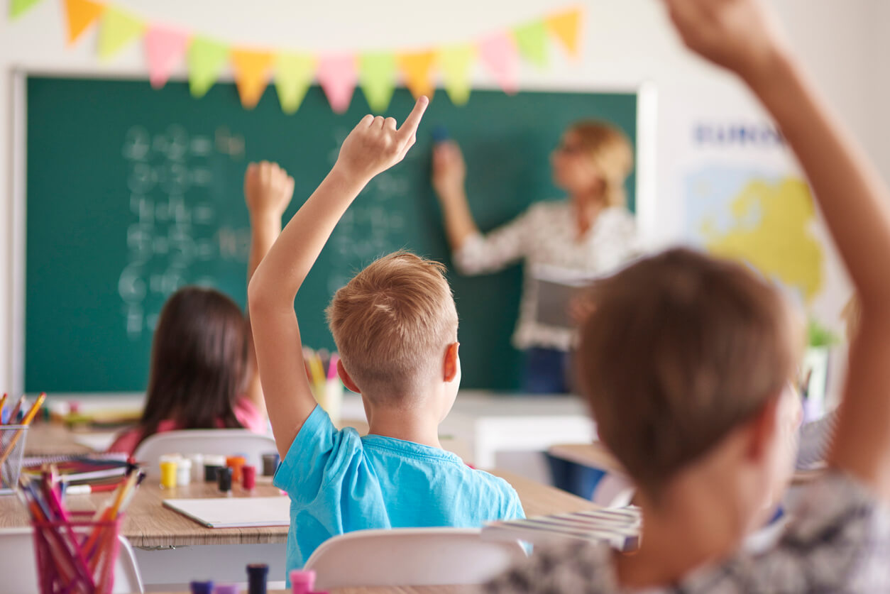 Elementary children raising their hands while their teacher teaches math on the chalkboard.