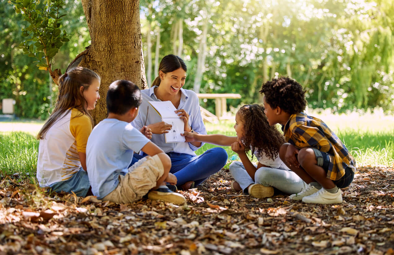 Children sitting under a tree with an adult who's holding a notebook with a drawing on it.