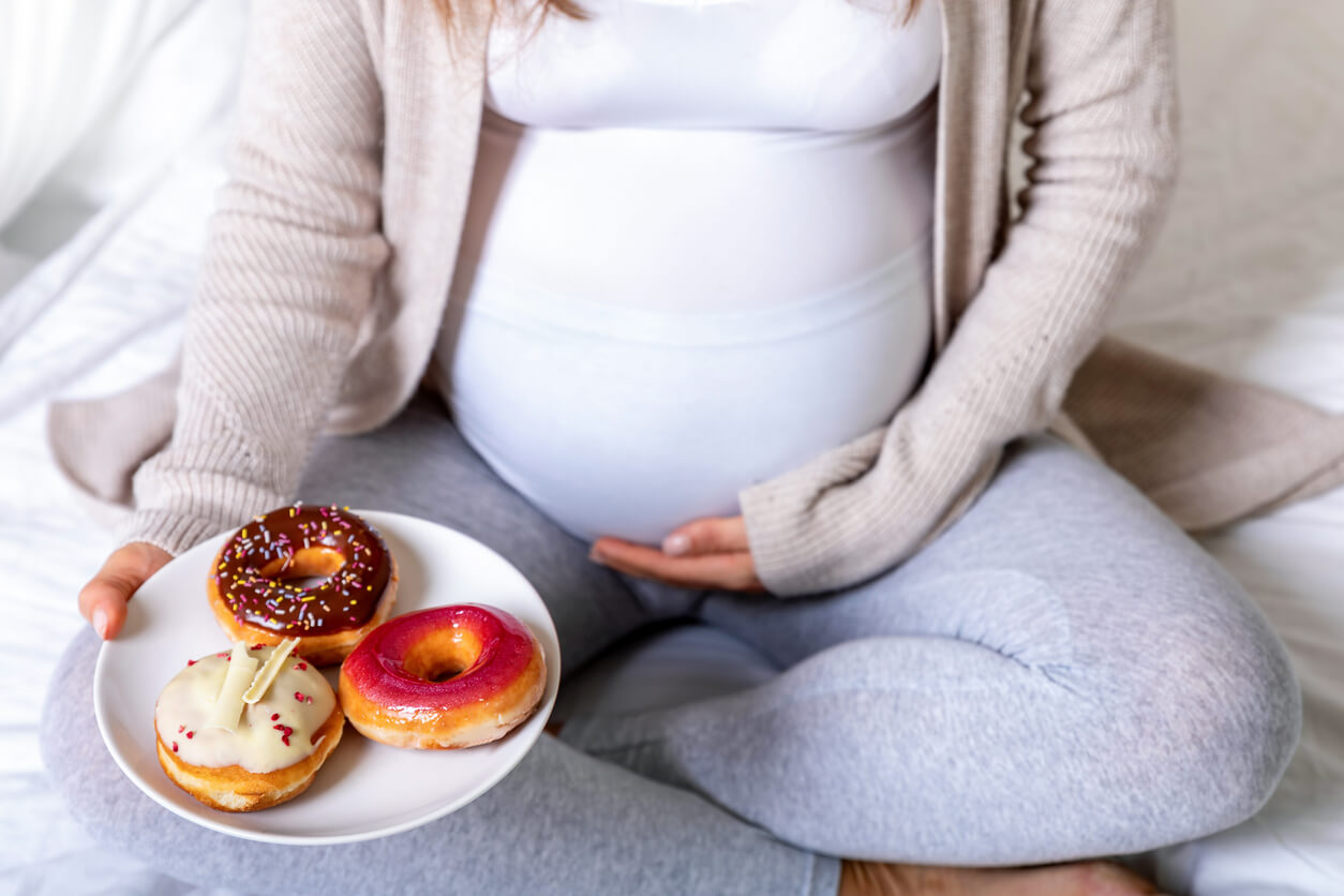 A pregnant woman holding a plate with three frosted donuts on it.