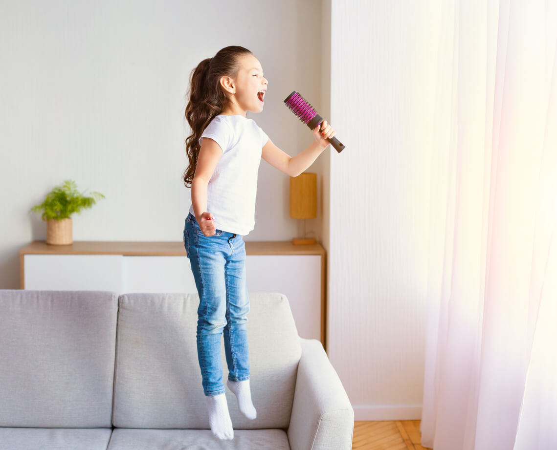 A young girl jumping on the couch and singing while susing a hair brush as a microphone.