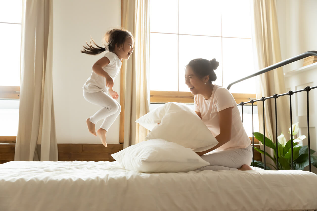 An Asian mother laughing while her daughter jumps on the bed.