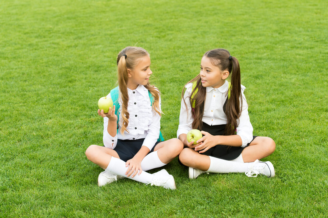 Two boarding school girls sitting in the grass talking and eating apples.