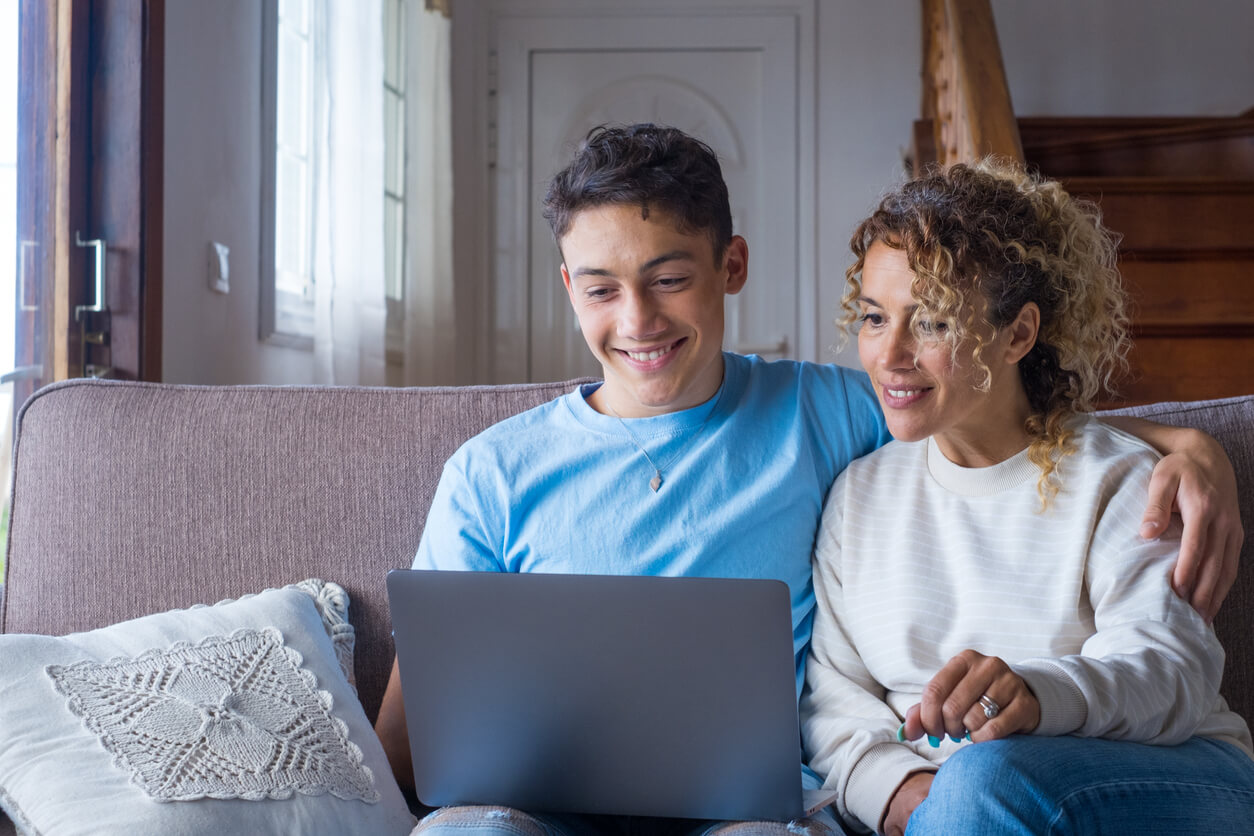 A mother and her teenage son sitting on the couch together, looking at a laptop screen.