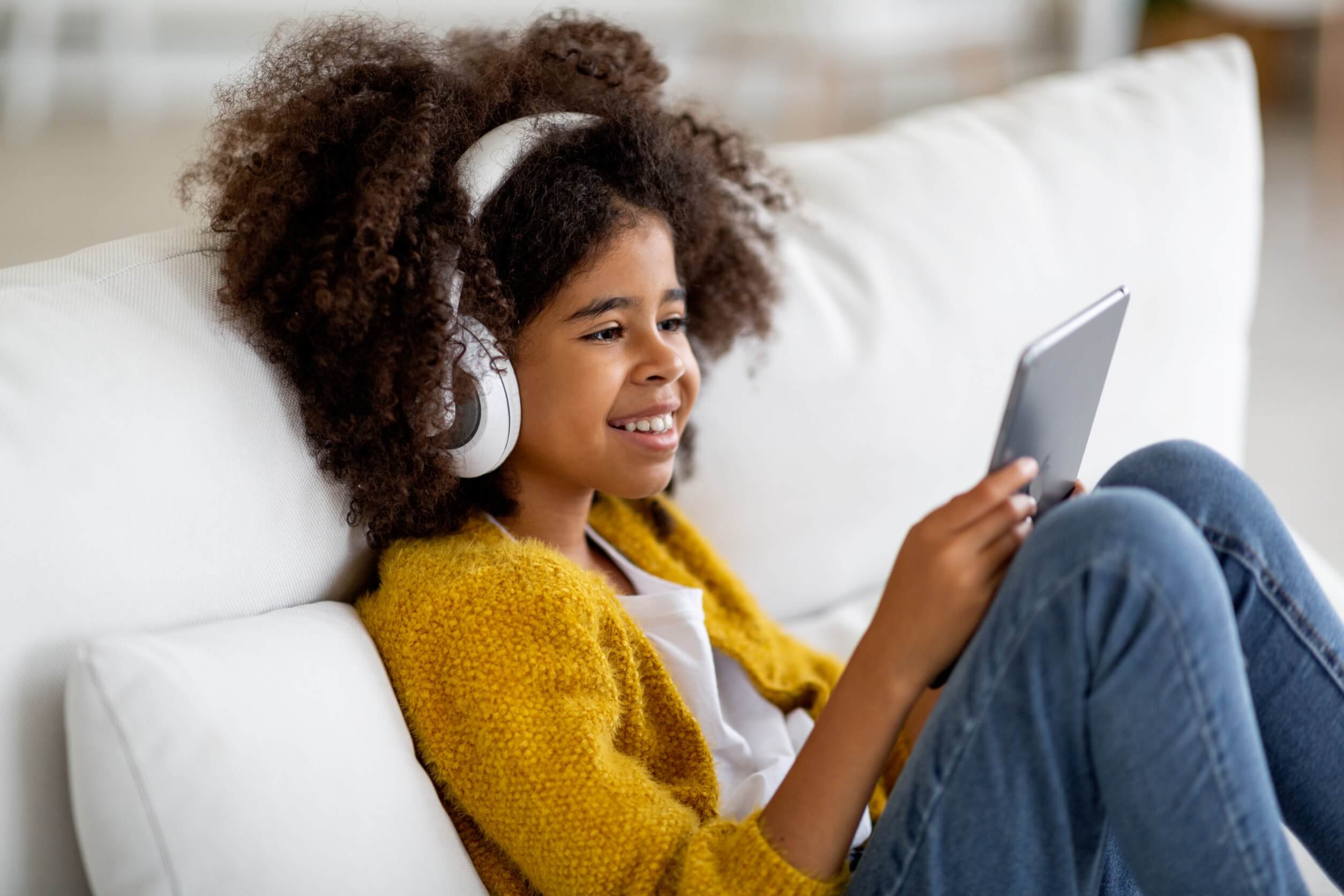 A little girl wearing headphones and watching a tablet, smiling.