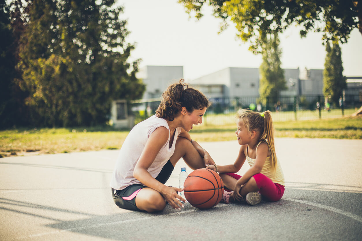 A mother encouraging her daughter while sitting on a basketball court in a park.