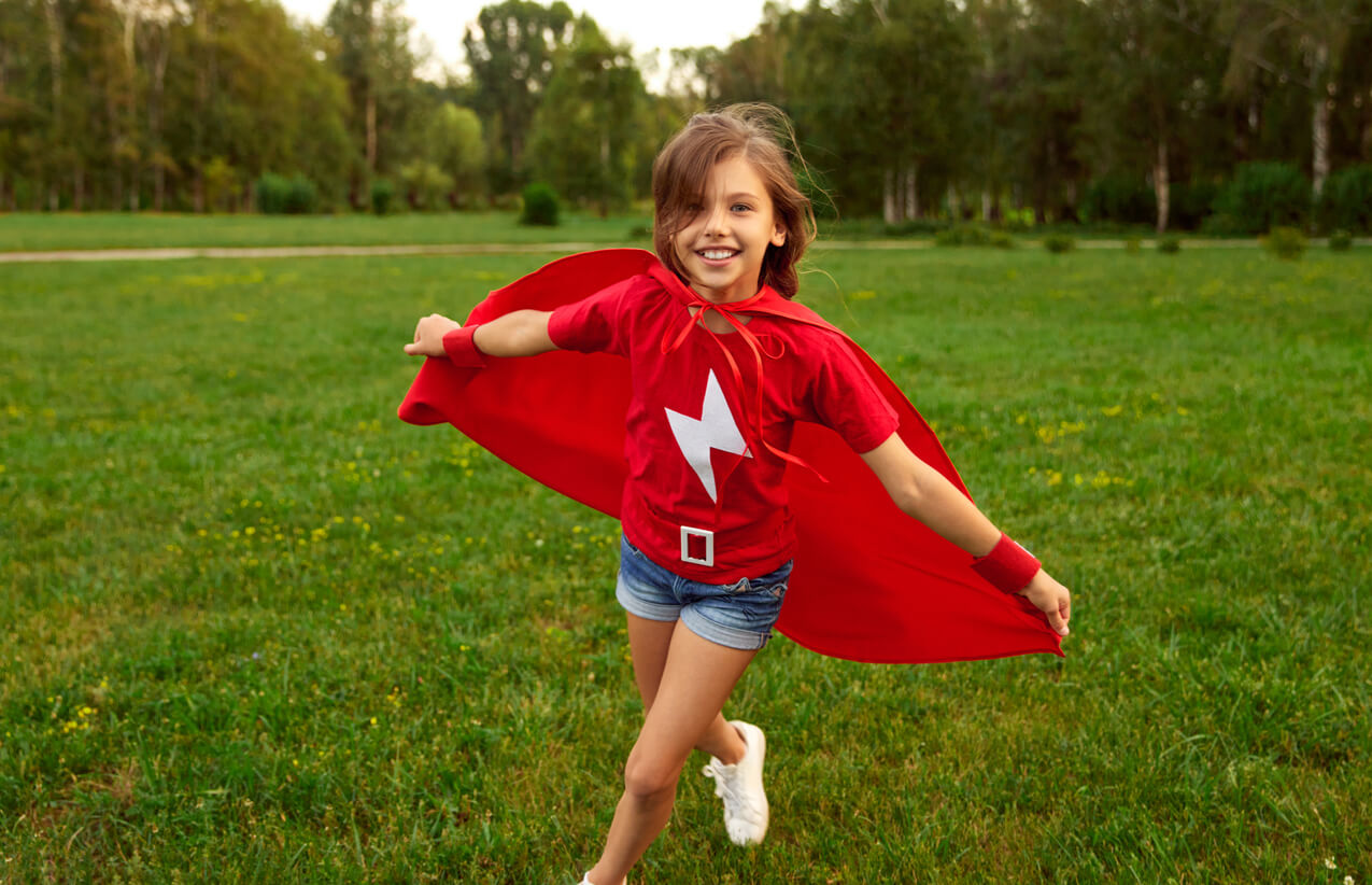 A girl running in the park with a superhero costume on, smiling.