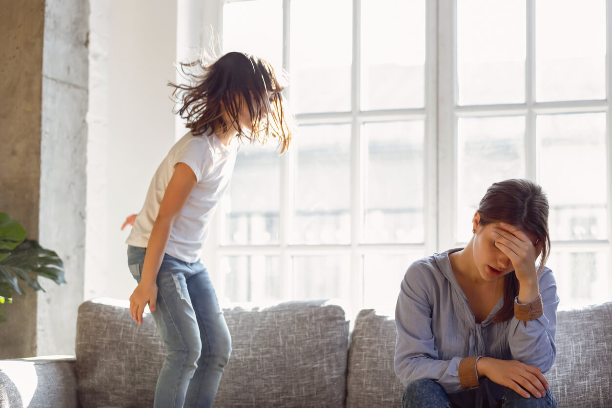 Une maman fatiguée à côté de son enfant qui saute sur le canapé.