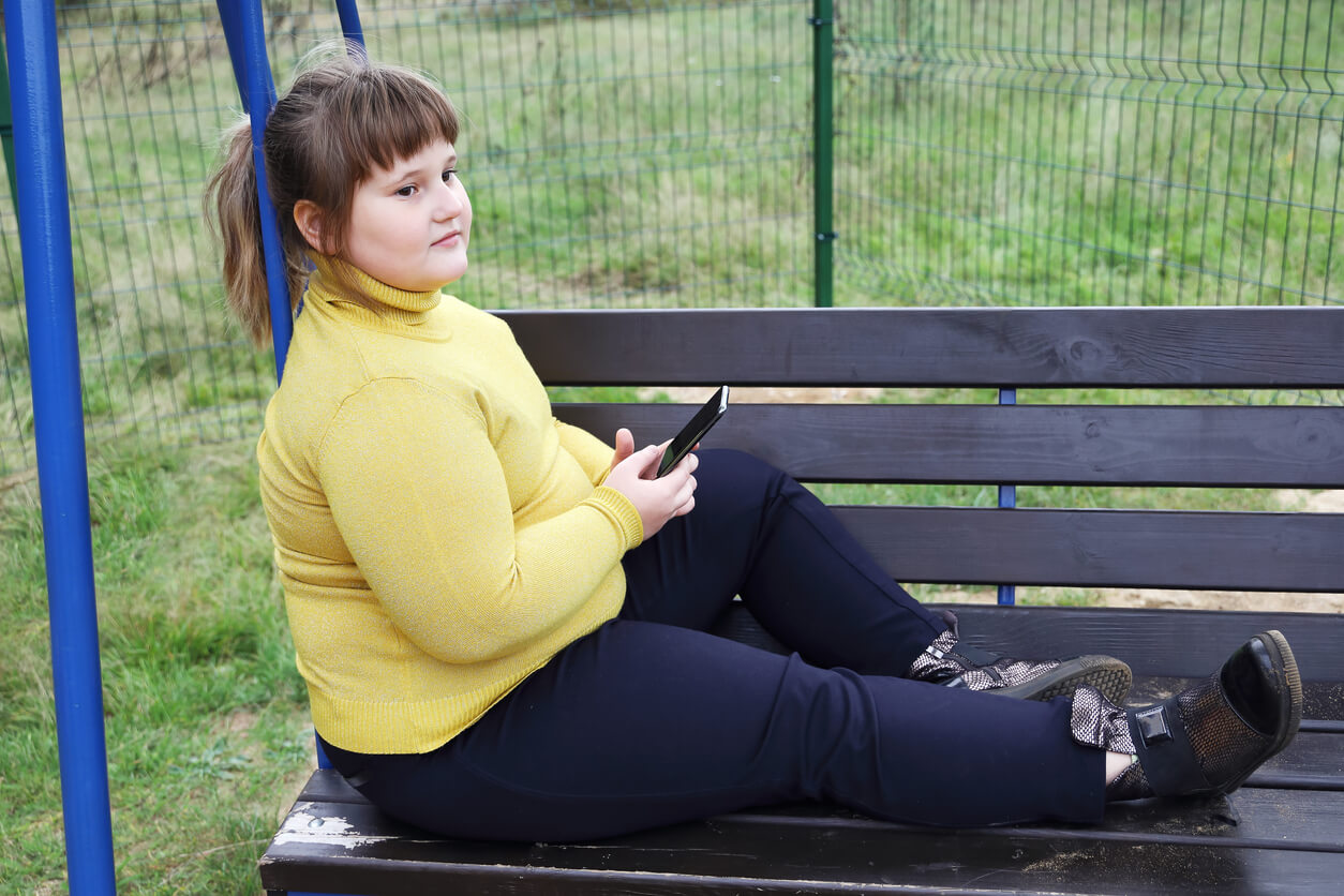 Ylipainoinen lapsi istuu puiston penkillä ja katselee matkapuhelinta.