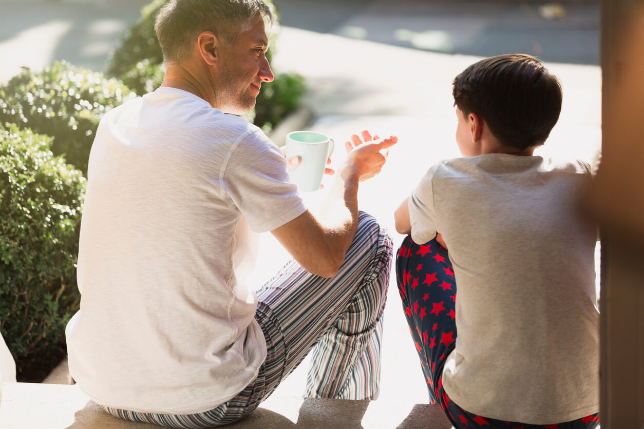 A father and son sitting down and talking on the porch in their pajamas.