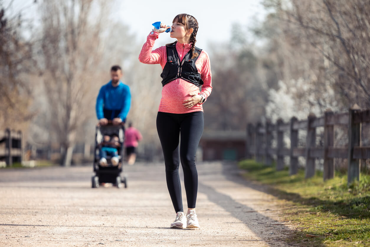 A pregnant woman walking in the park and drinking water.