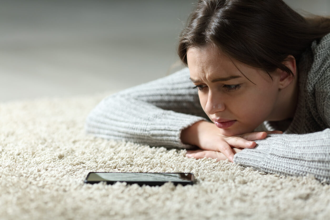 A teen girl lying on the floor, looking at her cell phone and crying.