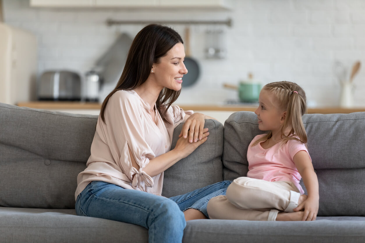 A mother and her young daughter sitting on the couch smiling and talking.