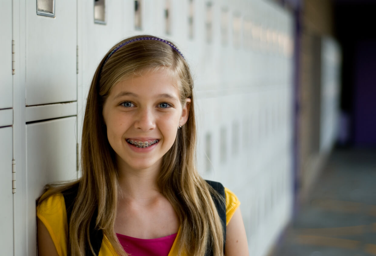 A young teen girl with braces standing by a row of school lockers.
