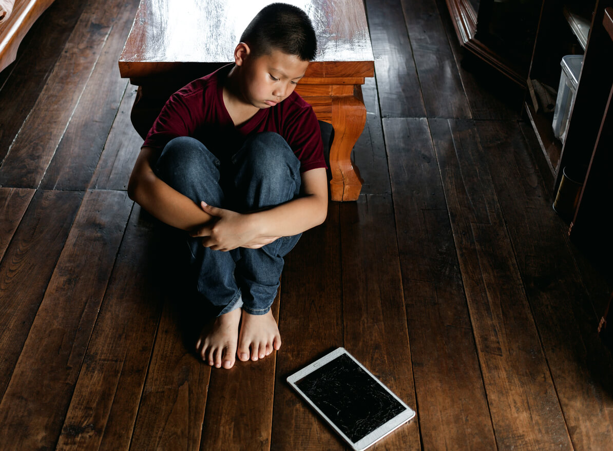 An Asian pre-teen boy looking unhappily at a tablet lying on the floor.