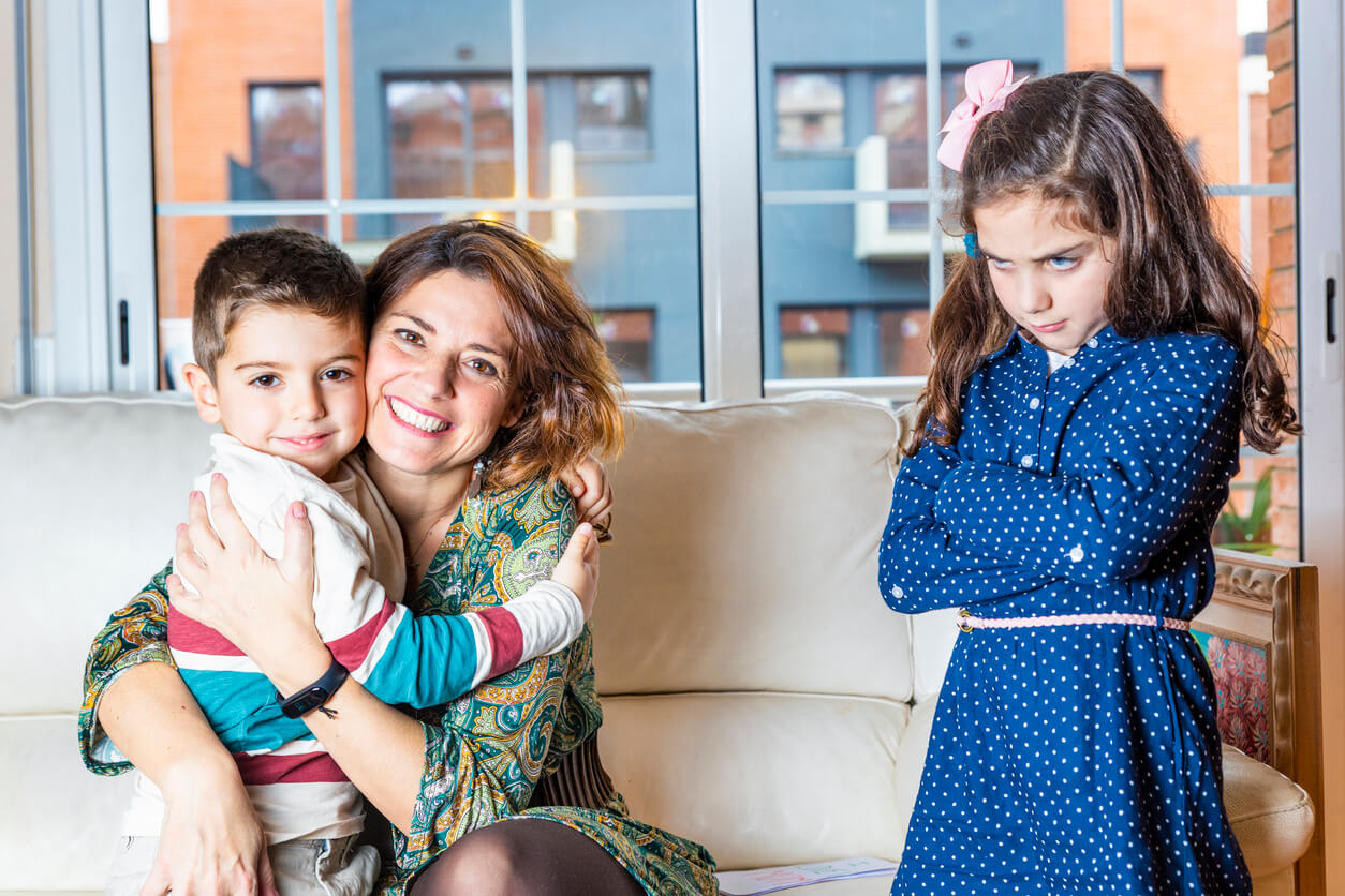 A mother hugging her young son while his big sister crosses her arm and pouts.