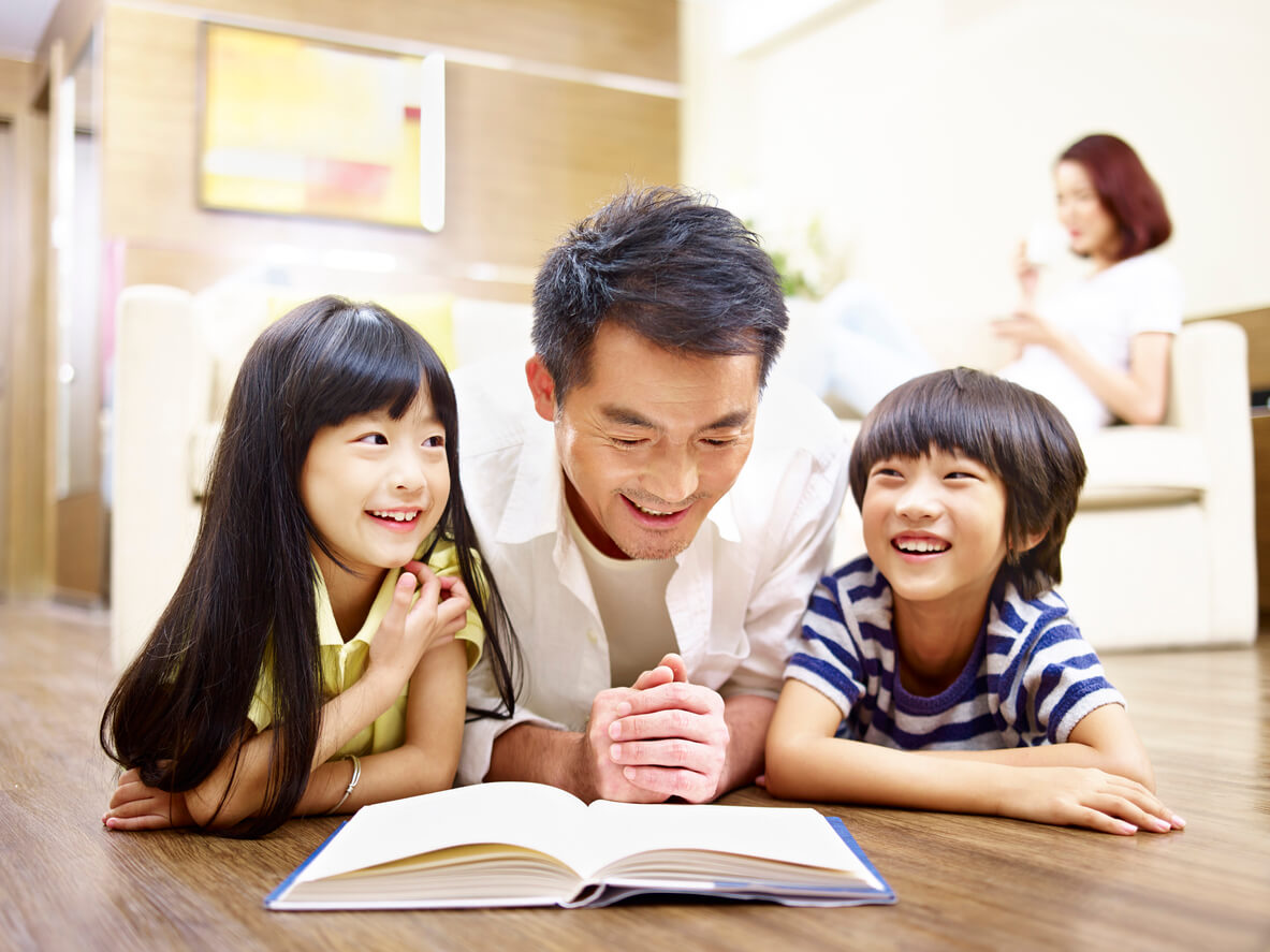An Asian father lying on the floor reading to his young son and daughter while their mother sits on the couch in the background drinking tea.