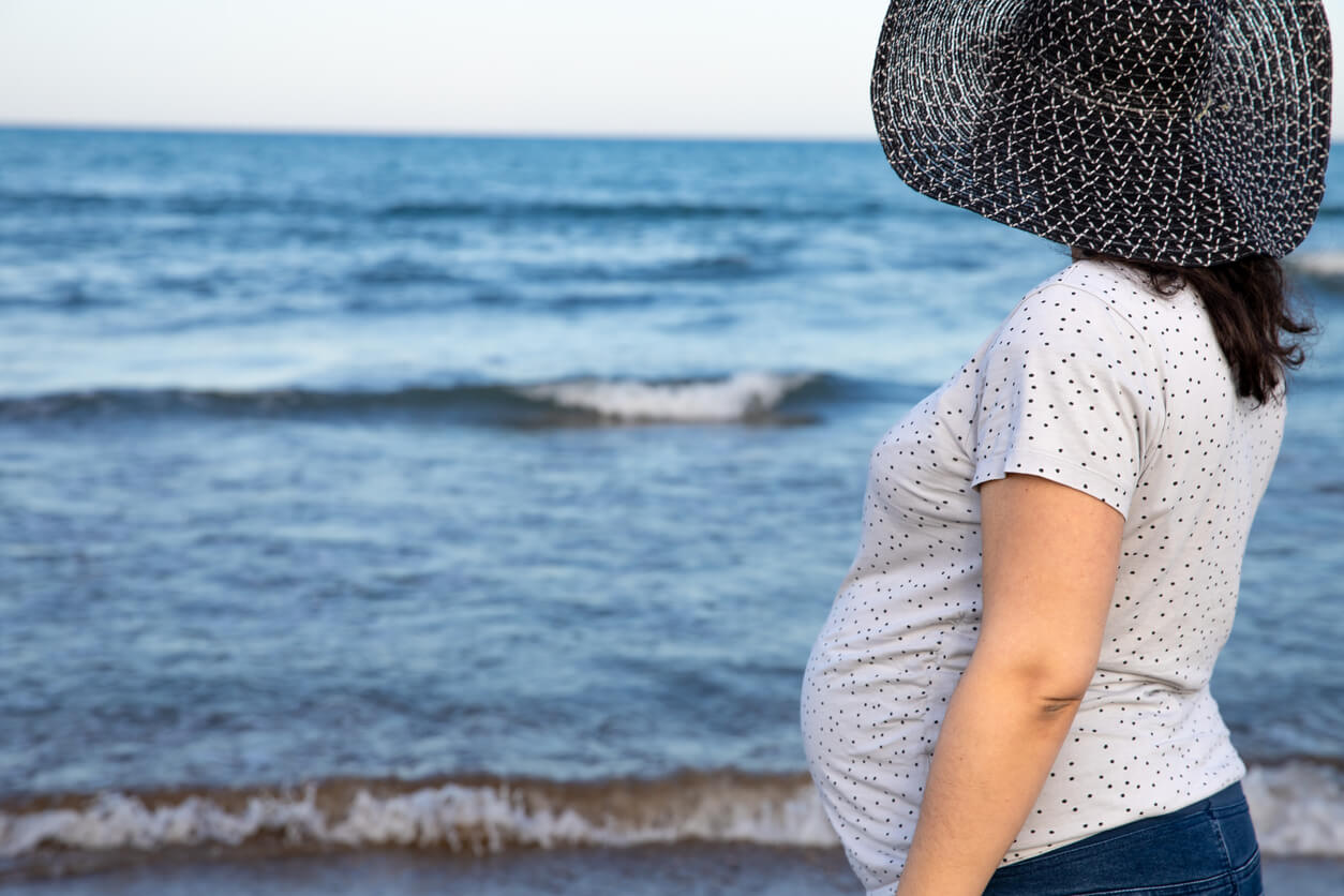 A pregnant woman wearing a wide-brimmed hat, looking out at the ocean. 