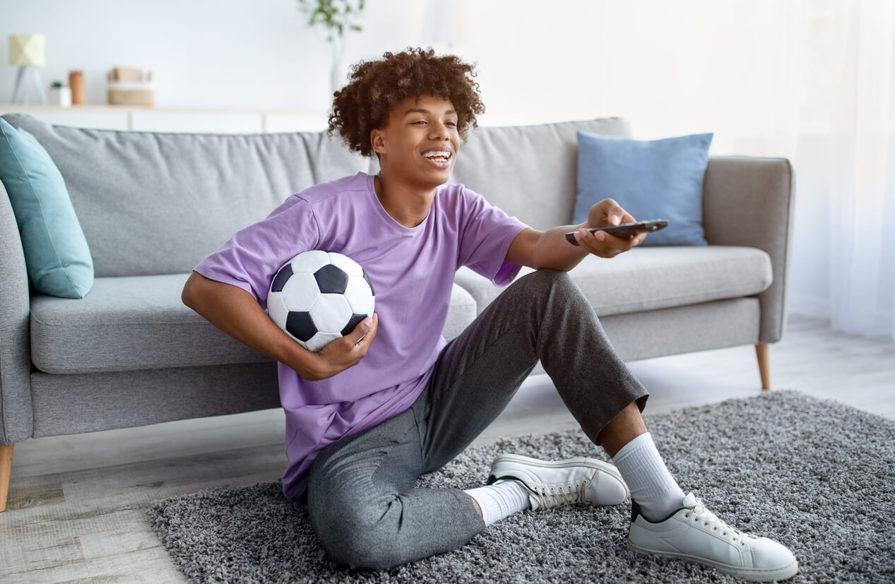 A black teenager sitting on the floor watching TV with a soccer ball under his arm.