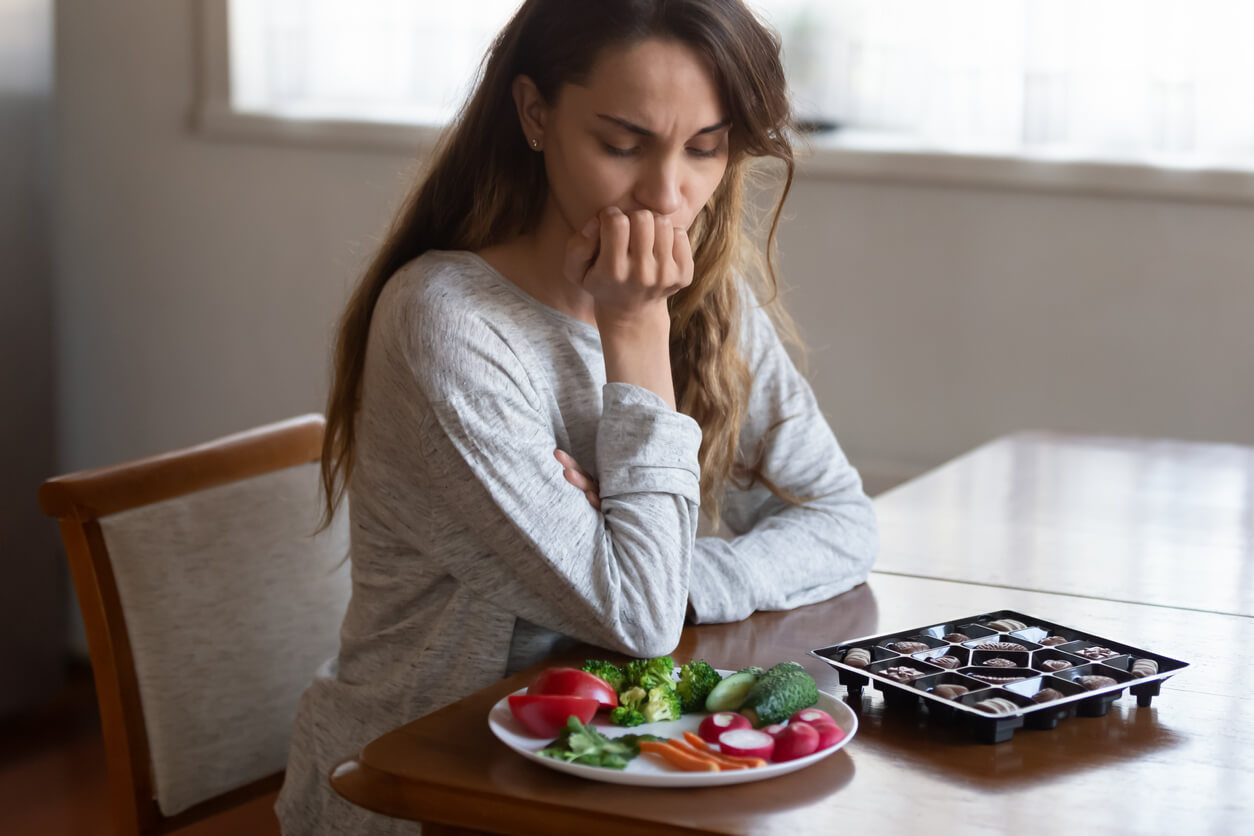 A woman staring at a plate of vegetables next to a box of chocolates.