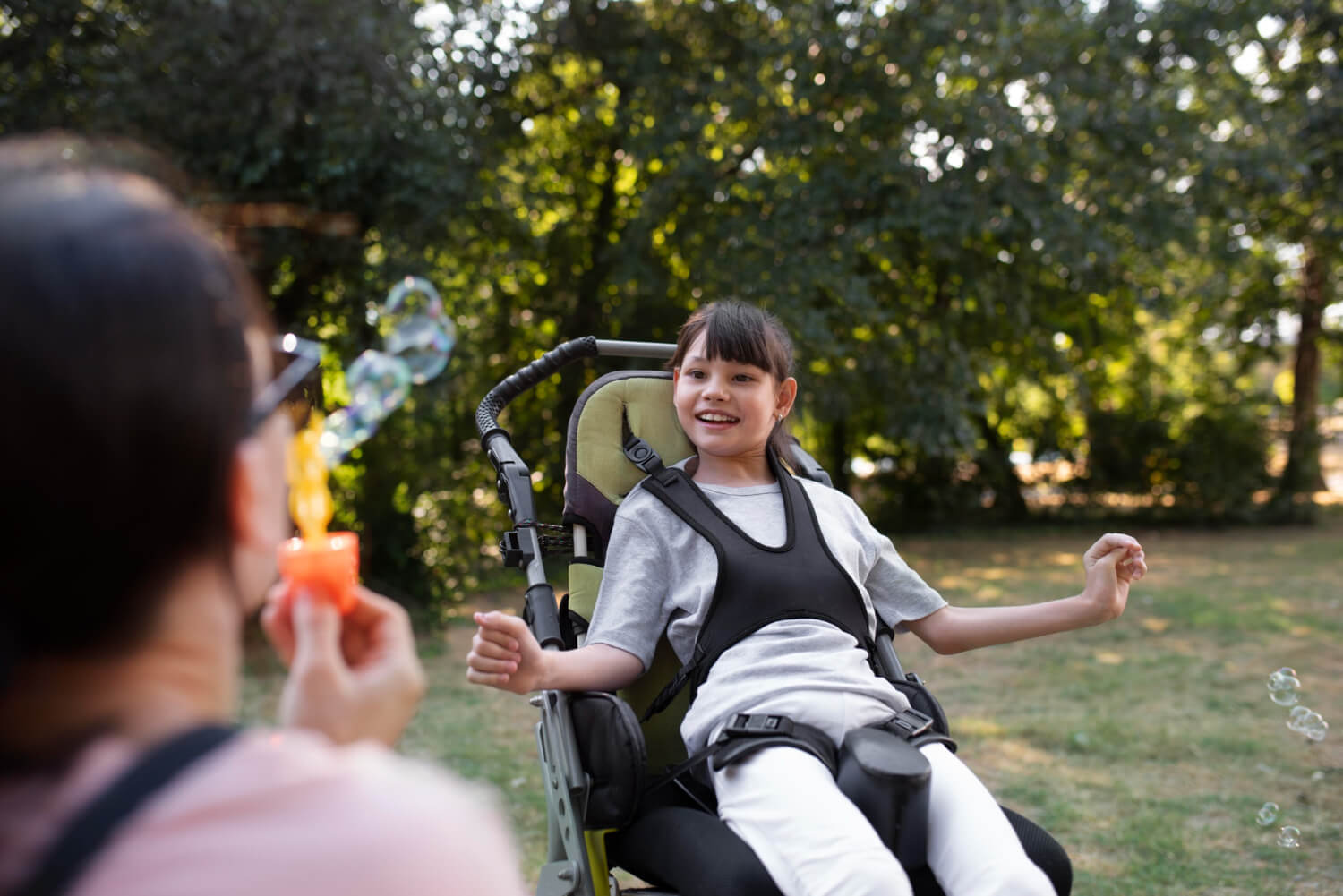 En preteen-jente med funksjonshemning smiler i rullestolen mens en annen person blåser såpebobler i luften.