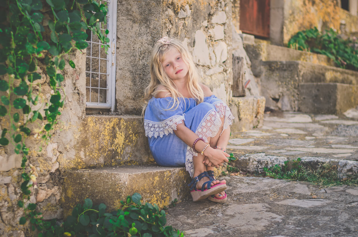A little blonde girl sitting on a doorstep in an old village.