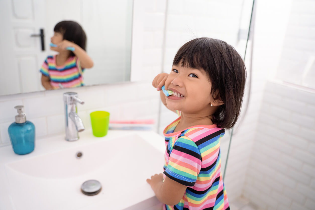An Asian toddler brushing her teeth and smiling.
