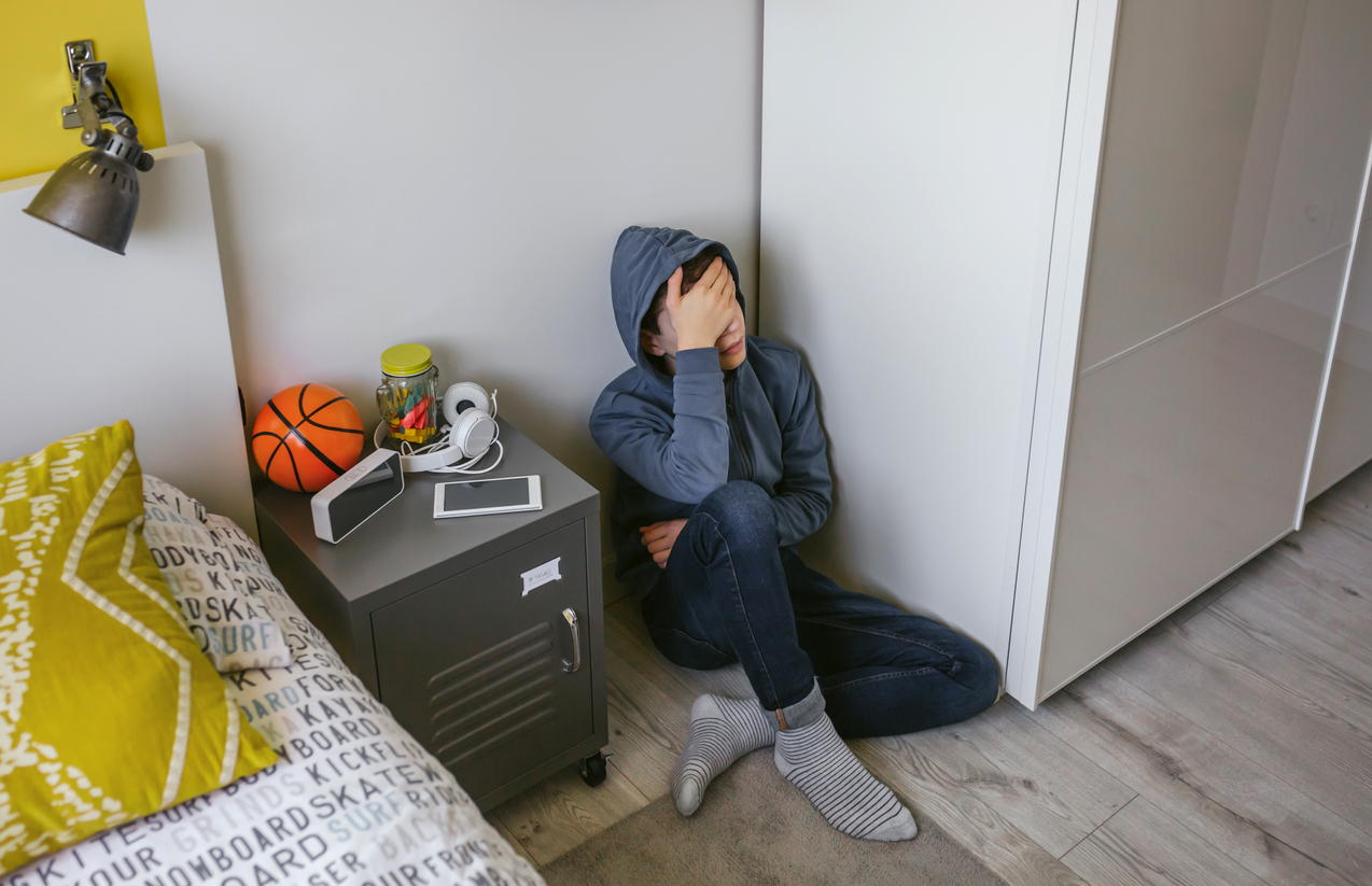 A teenager sitting on his bedroom floor, covering his face with his hand.