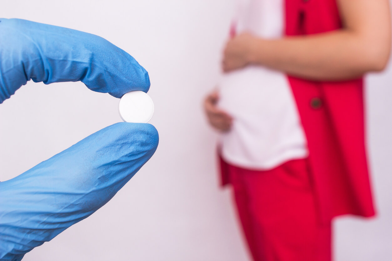 A gloved hand holding a pill in the foreground, with a pregnant woman standing in the background.