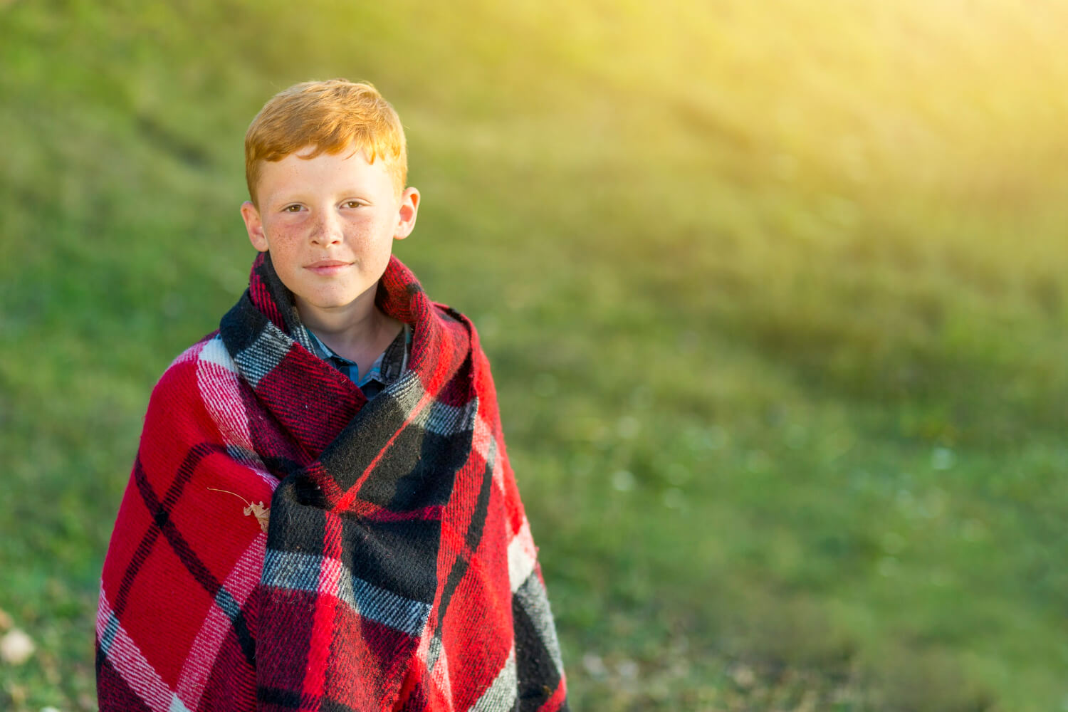 A red-headed boy wrapped in a red plaid blanket, standing on a grassy hill.