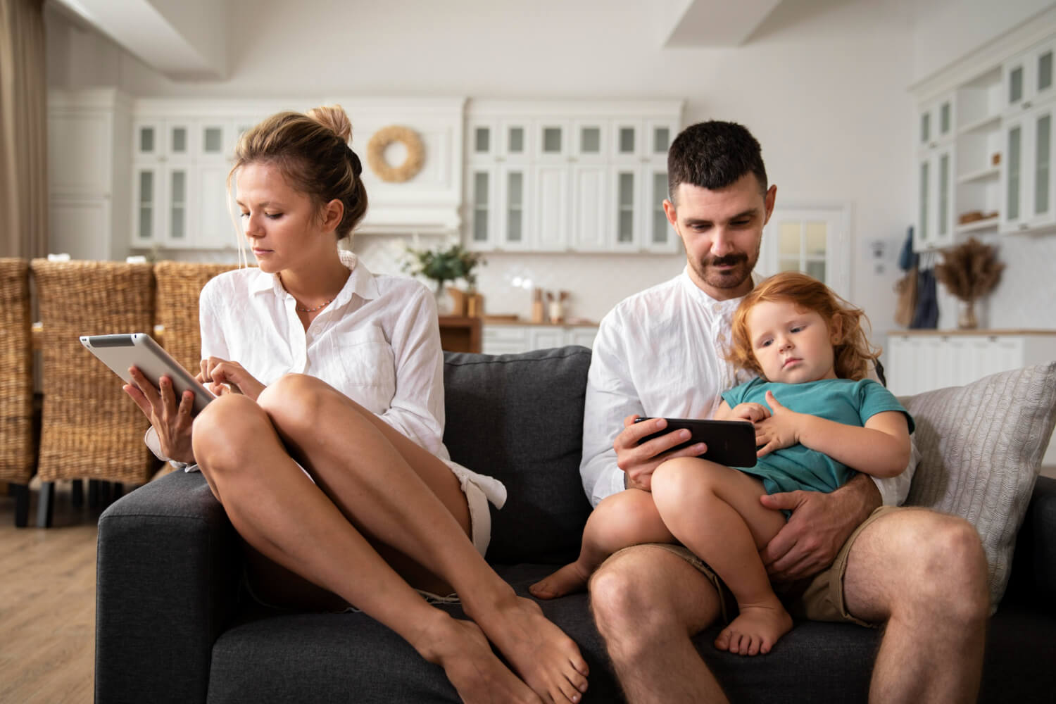 A woman looking at a tablet while her partner looks at a phone with his daughter.